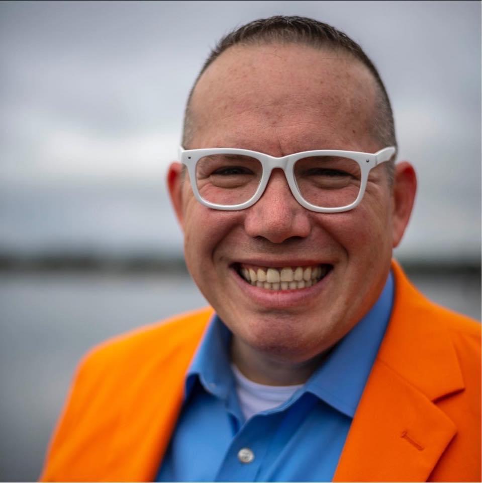 Profile photo of Phil Gerbyshak in blurred background wearing and bright orange jacket and blue shirt with white rimmed glasses